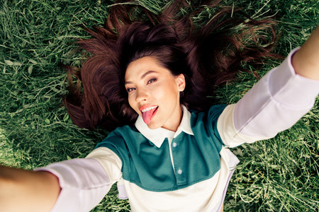 Positive woman enjoying outdoor leisure time, lying on green grass while making a playful selfie...