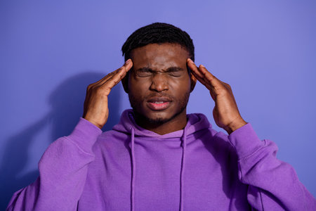 Young African American man in purple hoodie touching temples against purple background conveying...