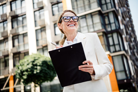 Charming businesswoman stands outdoors holding a clipboard, smiling under the sunshine with modern architectureの写真素材