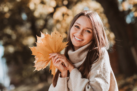 Charming young woman in stylish casual autumn wear holding colorful leaves in park enjoying outdoor...