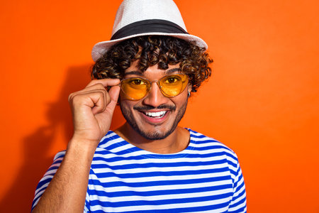 Cheerful young man in striped shirt with sunglass and hat, posing against bright orange backdrop,...