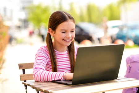 Smiling young girl using a laptop outdoors on a sunny day in a casual environment with natural light