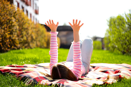Child relaxing outdoors on a sunny day, raising hands with joy while lying on a blanket in a green...