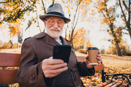 Elderly man enjoys outdoor autumn day holding coffee and phone in serene city park with vibrant...