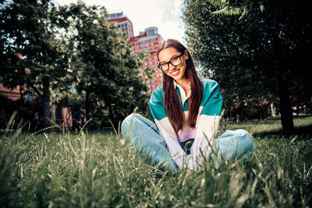 Smiling young woman sitting on grass in a city park dressed in casual style enjoying a sunny day...