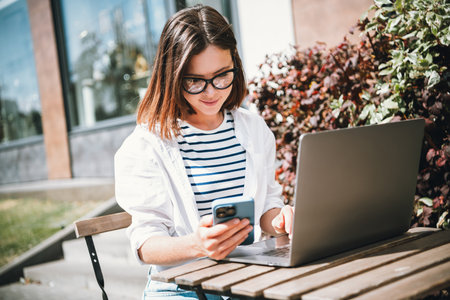 Young woman enjoying a sunny day outdoors, connected on phone and laptop, wearing stylish casual...