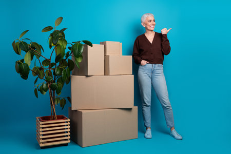 Happy mature woman with short gray hair posing with cardboard boxes and a plant on a blue background