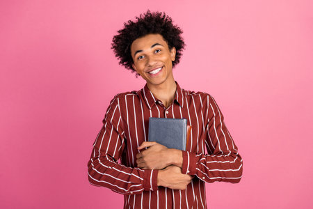 Smiling young man holding a book in a striped shirt against a pink background