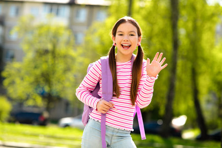 Happy schoolgirl waving with a smile while standing outdoors in a sunny park