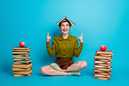 Young woman with book on head sitting on floor surrounded by colorful book and apples on blue...