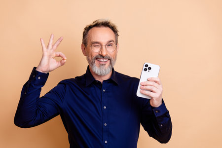 Mature businessman in elegant attire presenting smartphone against beige background