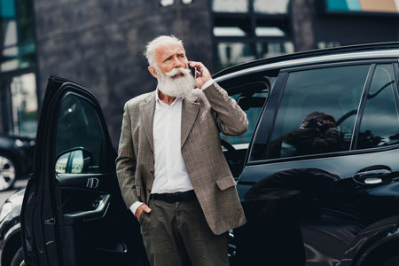 Confident mature businessman with grey beard making a phone call outdoors near his elegant car in...
