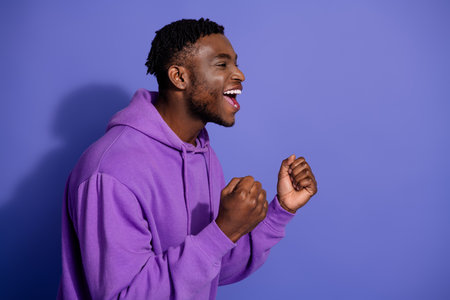 Excited young man wearing a purple hoodie, celebrating with enthusiasm against a solid violet...