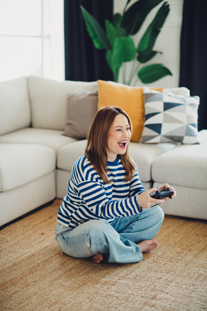 Woman sitting on floor in cozy living room playing video game with a controller, expressing joy and...