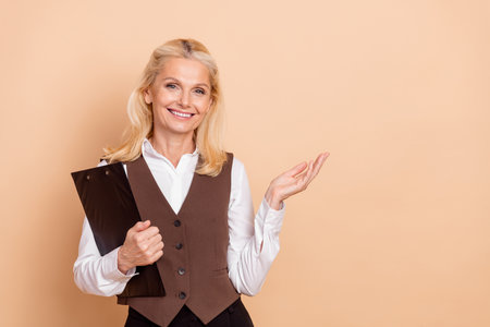 Elegant businesswoman wearing formal attire holding clipboard against beige background representing confident professional attitudeの写真素材