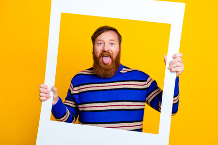 Young bearded man in a striped sweater posing playfully with a frame against a vibrant yellow...