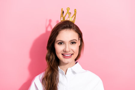 Charming young woman in white shirt wearing gold crown on a vibrant pink background showcasing...
