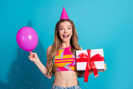 Cheerful young woman holding a gift box and a pink balloon against blue background, celebrating in...