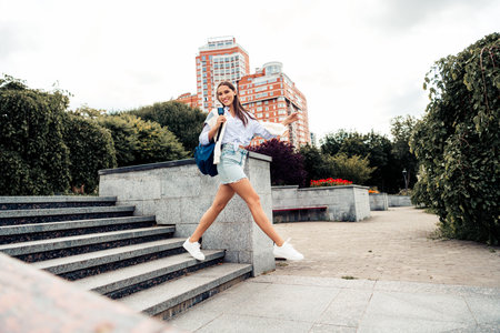 Young Stylish Woman Jumping Outdoors Near Urban Park and Modern Buildings in a Happy and Energetic...