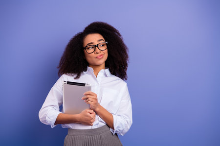Thoughtful young woman holding a tablet against a purple background, wearing glasses and formal attire, showcasing stylish confidenceの写真素材