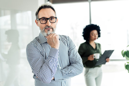 Confident businessman with a thoughtful expression standing in a modern office setting with diverse...