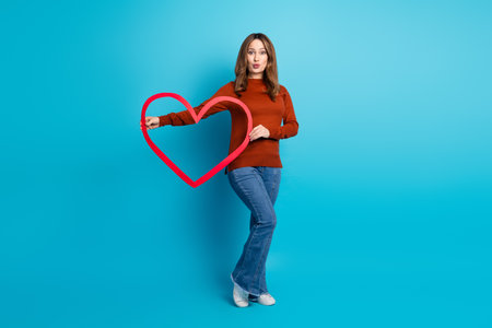 Charming woman holding a red heart in front of a blue background, expressing joy and charm with a casual styleの写真素材