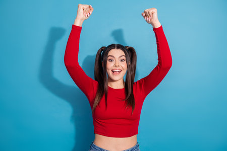 Excited Young Woman in Trendy Red Top with Upraised Hands Celebrates in Front of a Vibrant Blue...
