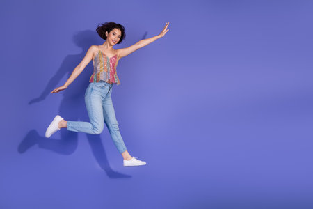 Cheerful woman in a stylish sequined top and jeans joyfully poses against vibrant violet background,...