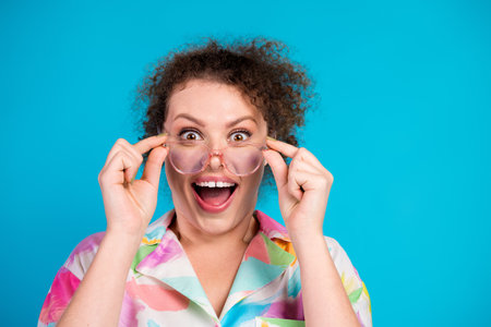 Excited young woman with curly hair in colorful shirt holding glasses in front of vibrant blue backgroundの写真素材