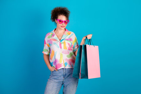 Stylish young woman with curly hair holding shopping bags in cheerful outfit against vibrant blue...