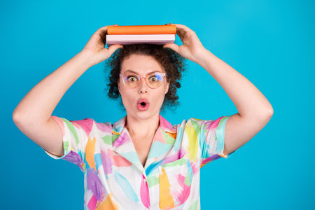 Playful woman with curly hair balances books on her head in colorful shirt against a bright blue...