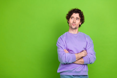 Handsome curly-haired man in purple sweater crosses arms while posing against vivid green studio...