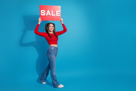Young woman holding a red sale sign against a vibrant blue background, expressing excitement and...