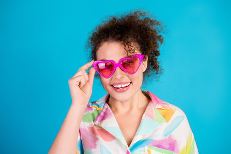 Cheerful young woman wearing pink heart-shaped sunglasses and colorful shirt posing with a bright...