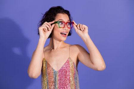 Young happy woman wearing colorful glasses and festive sequin dress, posing against a purple...