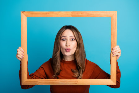 Portrait of a young woman holding a wooden frame expressing playfulness and creativity against a...