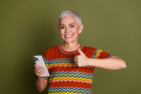 Happy senior woman with short grey hair holding a smartphone and giving a thumbs-up gesture against...