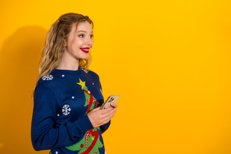 Young woman in Christmas sweater holding phone on festive yellow background, celebrating the holiday...