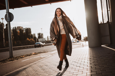 Young woman walking on an urban sidewalk in autumn, wearing a stylish coat, enjoying sunlight and...