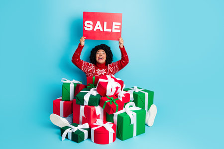 Festive woman in red Christmas sweater holding sale sign with colorful presents on a vibrant blue...