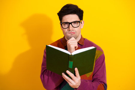 Young man in glasses reading a book thoughtfully against a yellow background, wearing a colorful...
