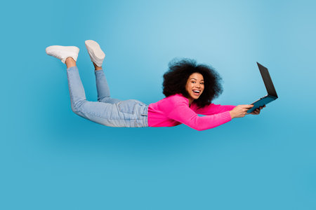 Young woman flying with a laptop on blue background, wearing casual outfit, displaying vibrant...