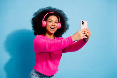 Happy young woman with afro hairstyle taking a selfie with smartphone wearing pink headphones...