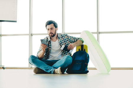 Man Sitting On Airport Floor Checking Phone While Traveling with Backpack and Personal Belongings