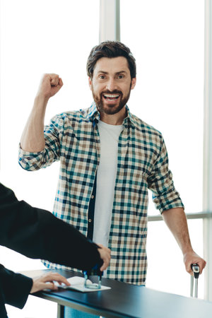 Excited man celebrating at airport terminal while holding suitcase near travel documents being stamped for boardingの写真素材