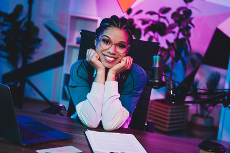 Young woman blogger in a neon-lit home office with microphone and computer preparing professional...