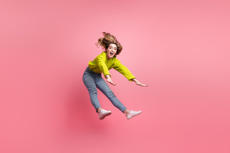 Adorable young woman in yellow sweater jumping with joy against a pink background showing cheerful...