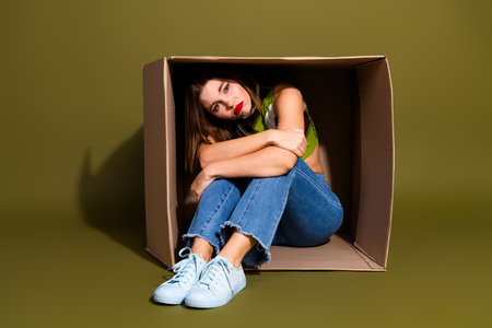 Stylish young woman sitting in a cardboard box against bright green background embodying casual...