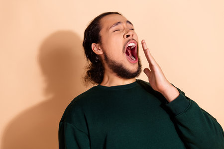 Portrait of a young man yawning while casually dressed, against a beige background, conveying...
