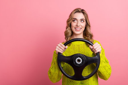 Adorable young woman holding a steering wheel, smiling joyfully against a vibrant pink background,...